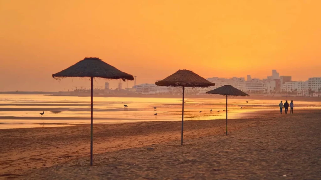 Golden sunset over Agadir's beach with silhouettes of straw parasols and people walking along calm waters.