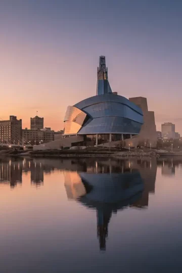 A panoramic shot of the Winnipeg skyline at sunset, featuring the curving glass architecture of the Canadian Museum for Human Rights and the Esplanade Riel bridge reflecting in the Red River.