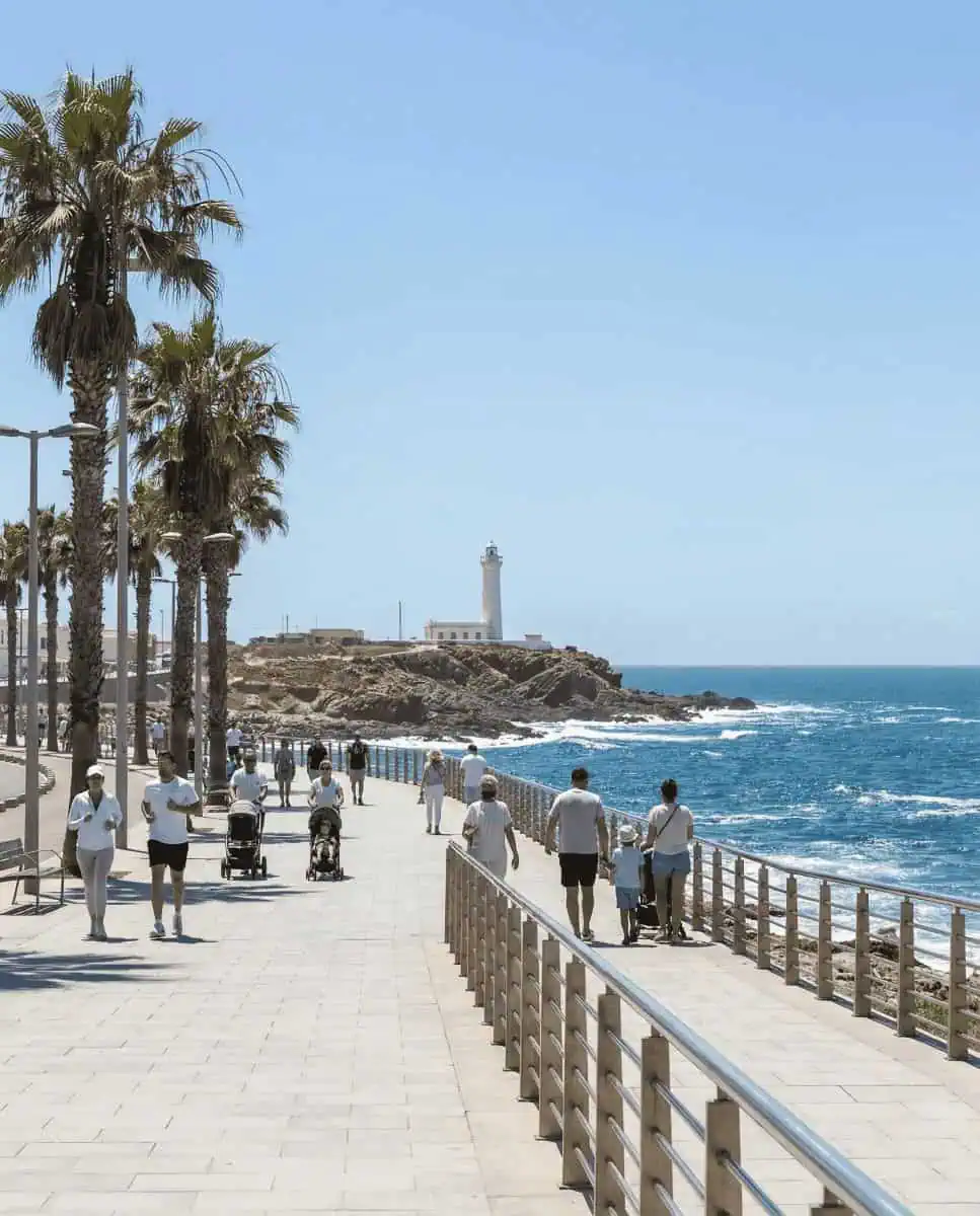 A bright, sunny view along the Corniche promenade looking towards the historic El Hank lighthouse standing on a rocky outcrop by the blue ocean.