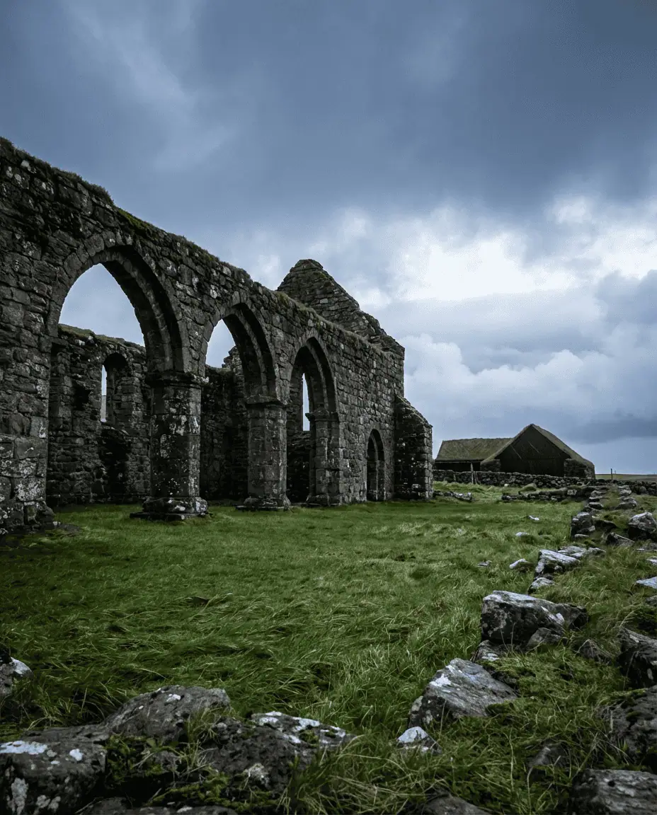 Low-angle shot of the stone ruins of Saint Magnus Cathedral in Kirkjubøur, with the black wooden farmhouse Roykstovan in the background under a dramatic sky