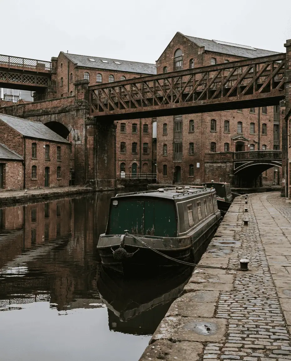 A peaceful daytime shot of the Castlefield canal basin, with a narrow boat moored by the towpath and the giant iron railway bridges towering overhead