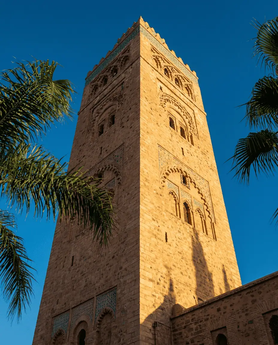 Low-angle shot of the towering Koutoubia Mosque minaret, its intricate stonework and ochre color contrasting sharply against a vivid deep blue sky, framed by swaying green palm trees.