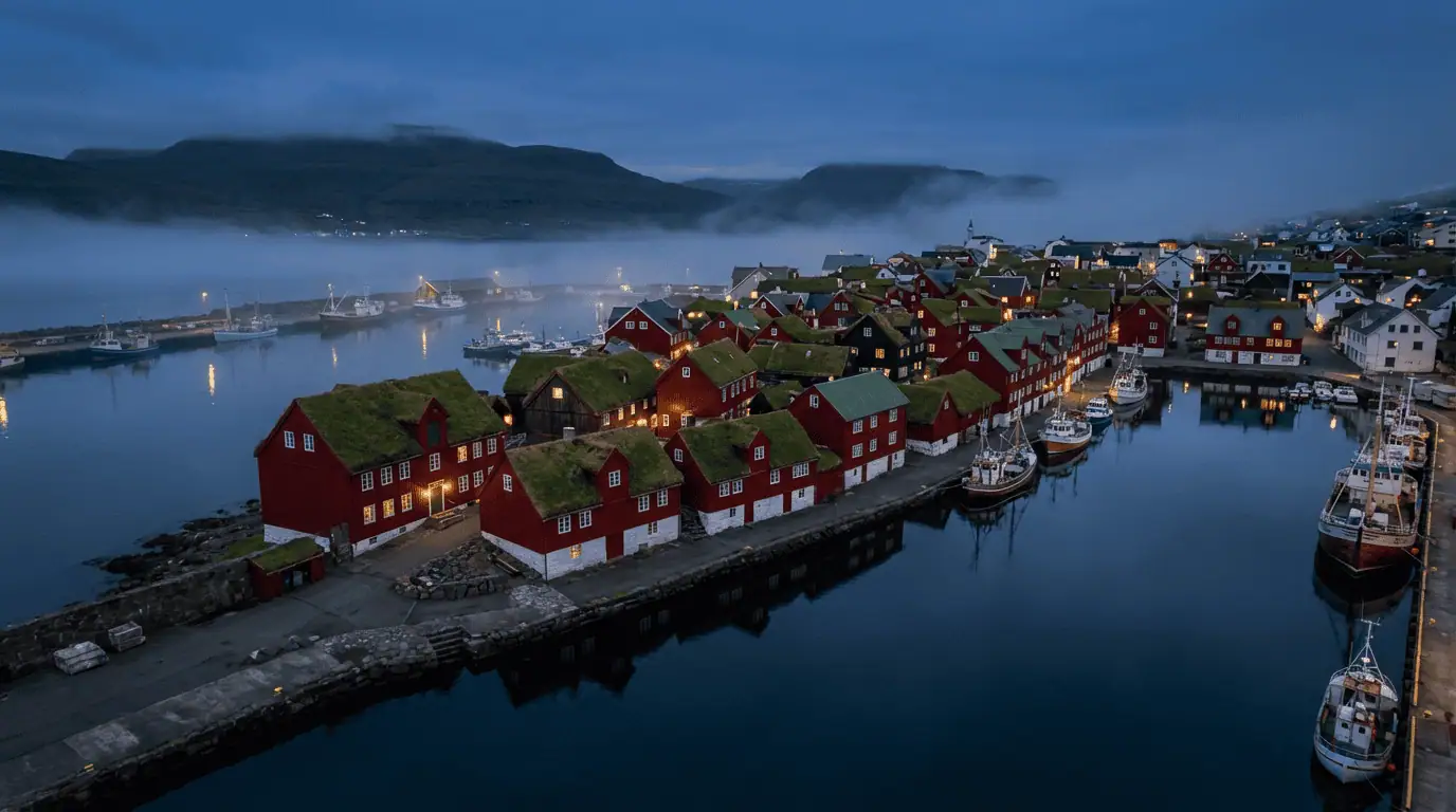 Aerial view of Tórshavn harbor at blue hour, showcasing the red turf-roofed buildings of Tinganes peninsula glowing against the deep blue North Atlantic ocean and moody fog-capped mountains.