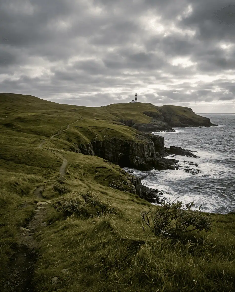 Wide landscape shot of the green hiking trail leading to the Nólsoy lighthouse, with the rugged coastline and crashing waves visible in the distance