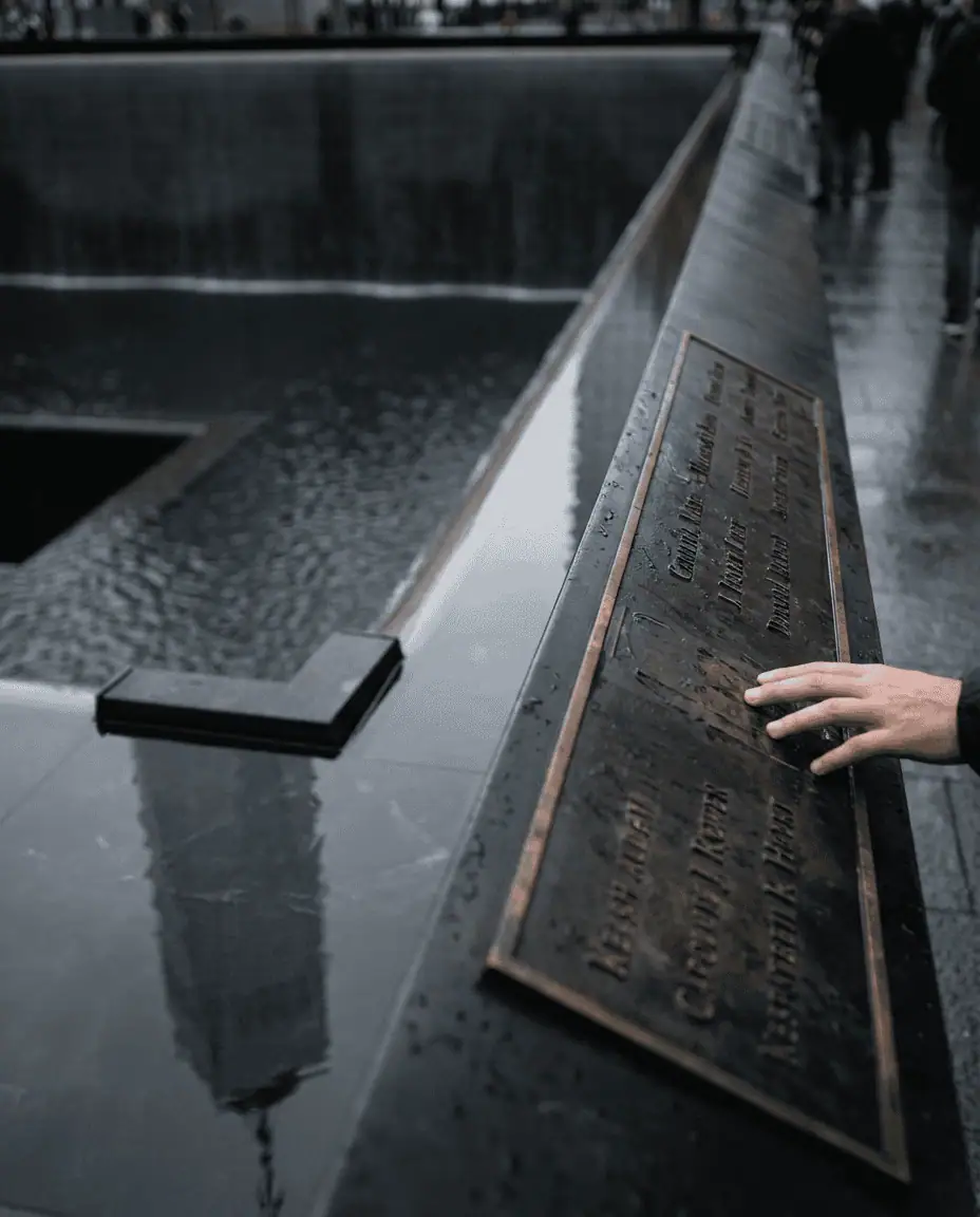 A somber, artistic photograph of one of the 9/11 Memorial reflecting pools, focusing on the bronze plaques inscribed with names, with the new World Trade Center tower reflecting on the water's surface.