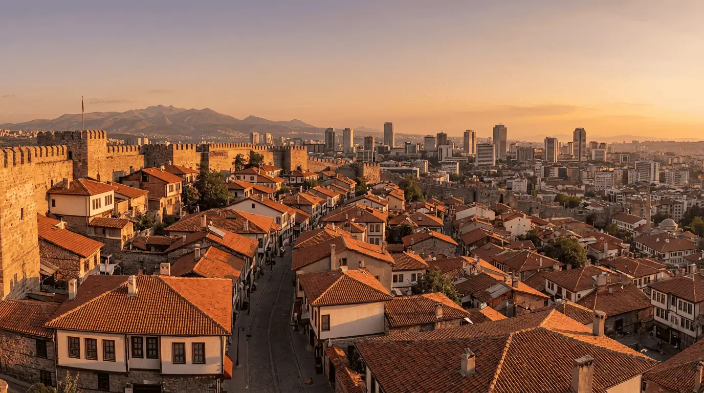A stunning wide-angle panoramic view from the top of Ankara Castle (Kale) during the golden hour, showing the red-tiled roofs of the historic old town in the foreground and the modern city skyline stretching to the mountains in the distance.