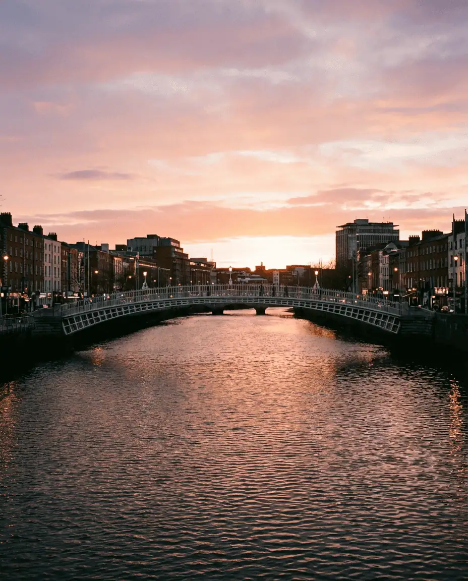 A romantic view of the white cast-iron Ha'penny Bridge spanning the River Liffey at sunset, with the city lights beginning to twinkle on the water.