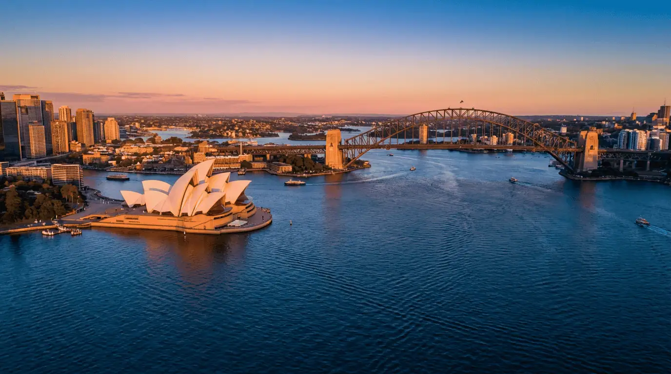 A stunning wide-angle aerial shot of Sydney Harbour at sunset, featuring the white sails of the Opera House and the steel arch of the Harbour Bridge glowing in golden light against a deep blue water background.