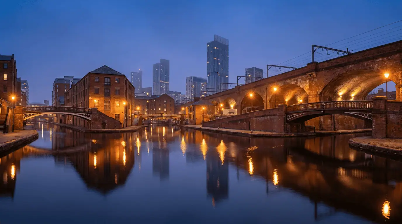 A panoramic view of the Castlefield canals at twilight, with the illuminated red-brick railway viaducts reflecting in the calm water and modern skyscrapers in the background.