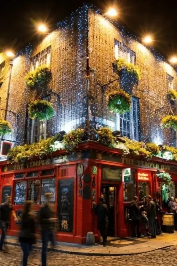 A vibrant, eye-level shot of the iconic red exterior of The Temple Bar pub, adorned with hanging flower baskets, on a bustling cobblestone street in Dublin's cultural quarter.