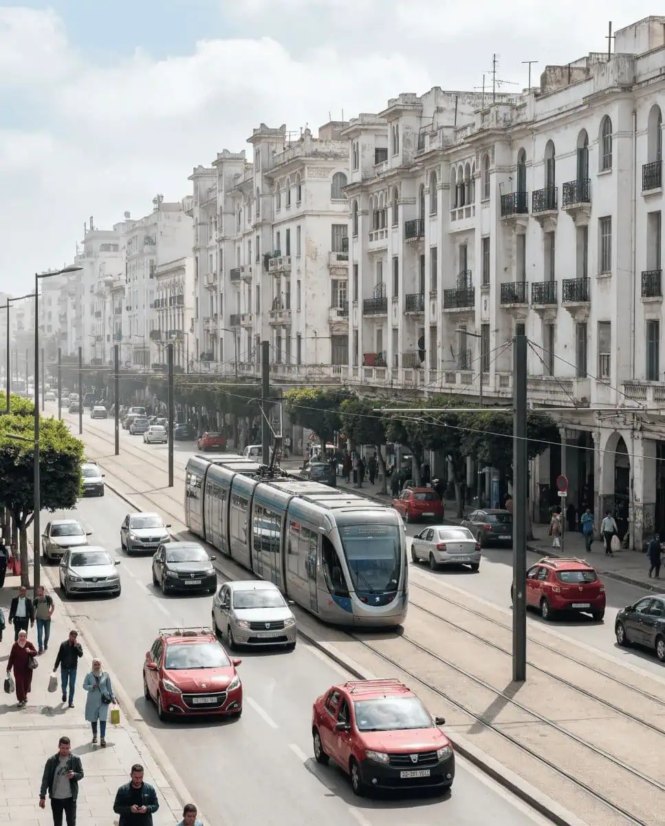 A dynamic street scene in downtown Casablanca showing a sleek modern tram passing by faded white Art Deco colonial buildings, illustrating the city's contrast.