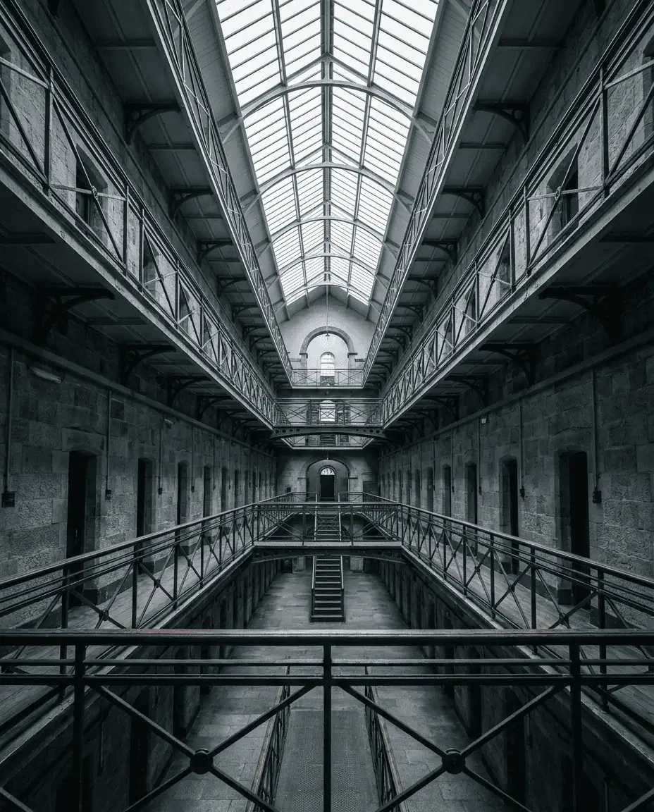 A dramatic, wide-angle interior shot of the Victorian East Wing of Kilmainham Gaol, showing the high metal walkways and the vast, empty central hall.